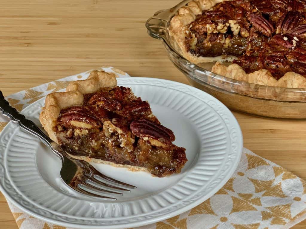 A slice of Pecan Pie with a thin chocolate layer served on a dessert plate beside the whole cut Pecan Pie.