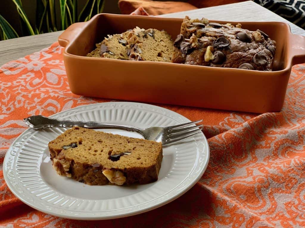 A loaf of Pumpkin Ribbon Bread, without the ribbon of cream cheese filling, topped with chocolate chips and toasted nuts beside a slice served on a plate.