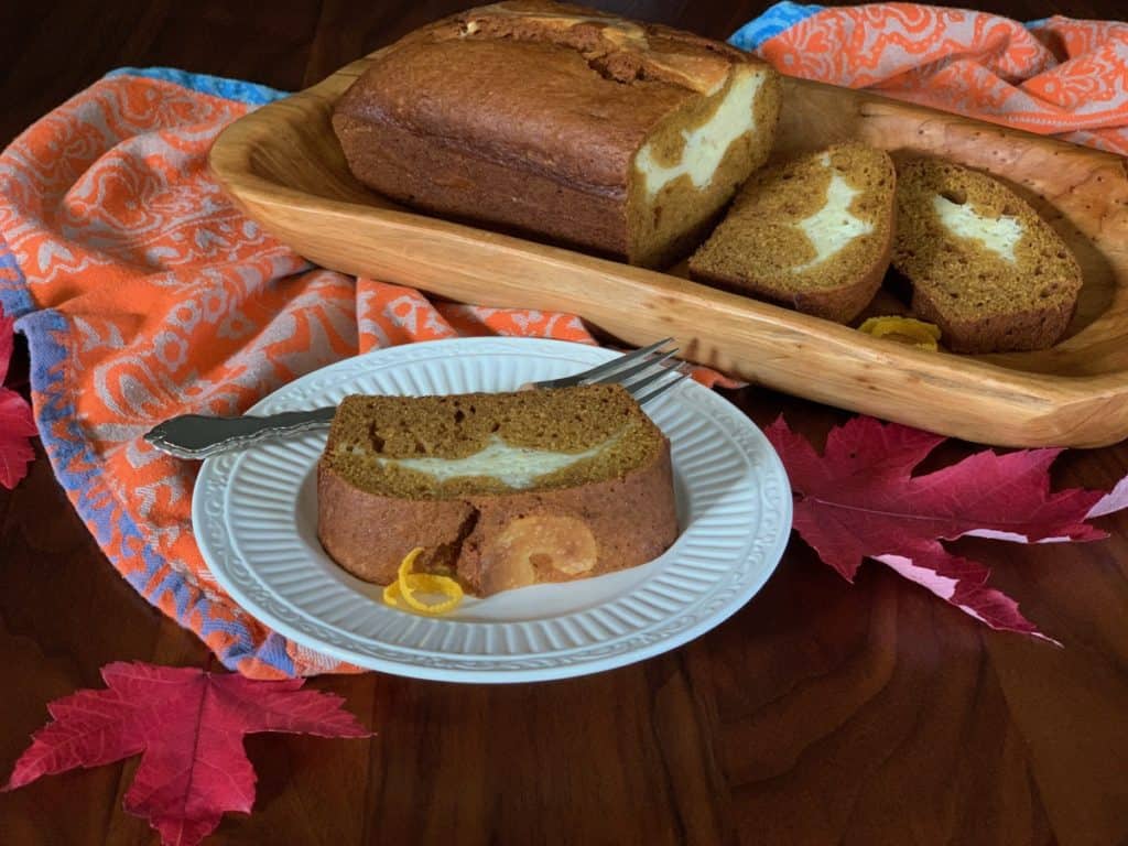 A sliced loaf of Pumpkin Ribbon Bread on a wooden tray beside a slice served on a white plate with a fork. 