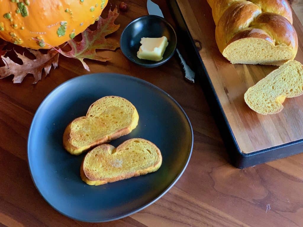 A Pumpkin Yeast Braid sliced on a wood cutting board with two slices toasted and buttered on a serving plate.