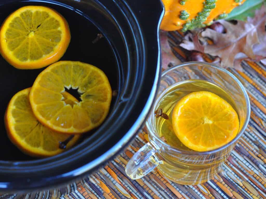 Mulled Cider with orange slices and cloves is served in a glass cup beside a ceramic pot filled with cider.