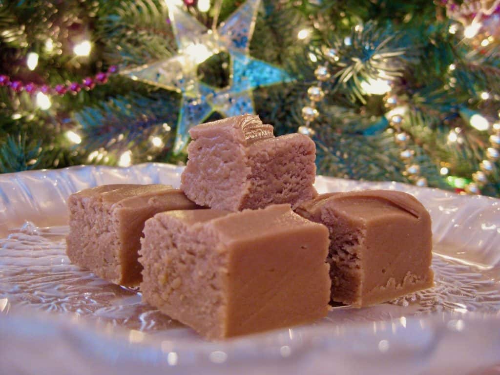 Squares of Peanut Butter Fudge stacked on a glass plate set in front of a lit Christmas tree.