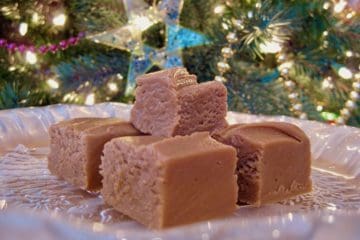 Squares of Peanut Butter Fudge stacked on a glass plate set in front of a lit Christmas tree.