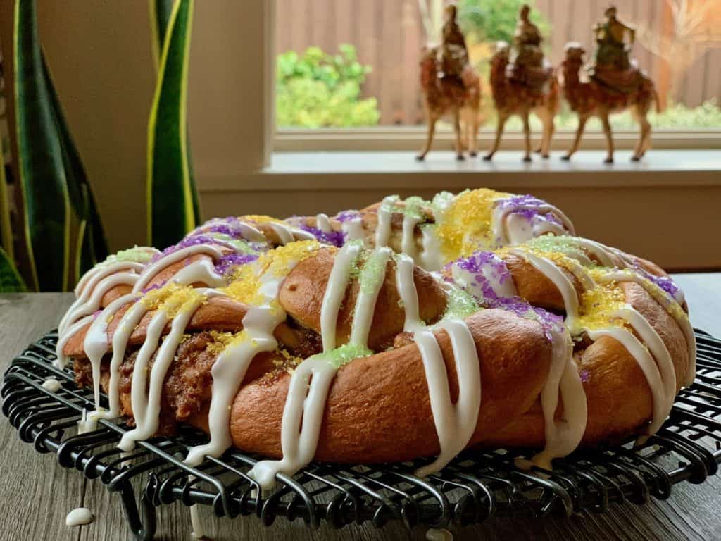 A braided King Cake drizzled with Vanilla Icing and sprinkled with Decorator Sugar rests on a wire rack with three kings on camels in the background. 