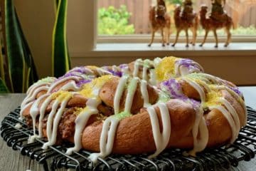 A braided King Cake drizzled with Vanilla Icing and sprinkled with Decorator Sugar rests on a wire rack with three kings on camels in the background.