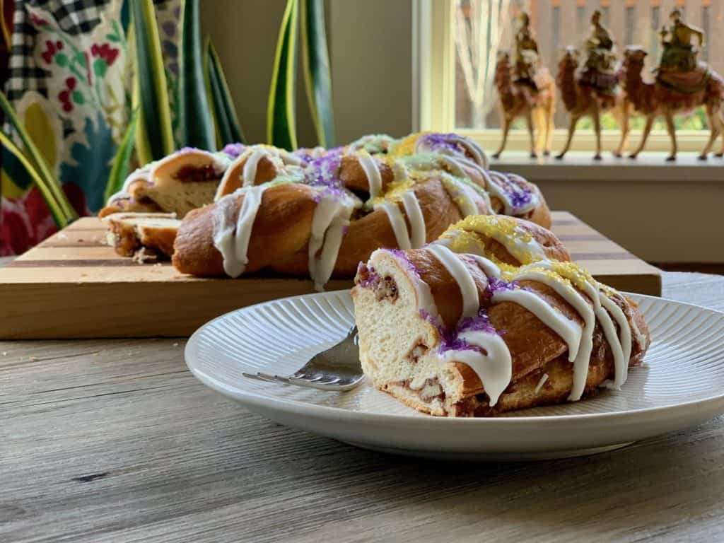 A braided King Cake drizzled with Vanilla Icing and sprinkled with Decorator Sugar sliced and served on a white dessert plate with three kings on camels in the background.