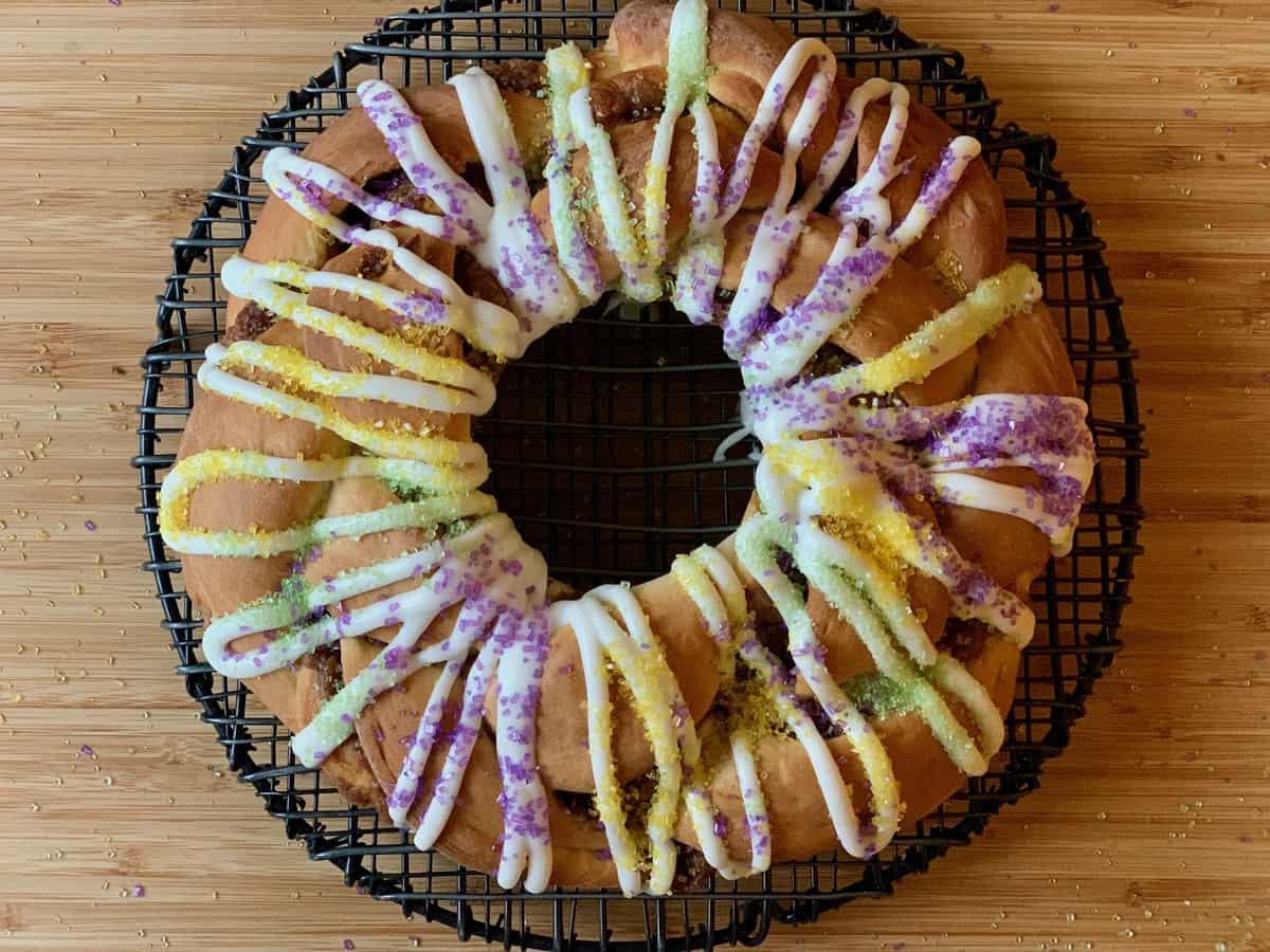 A braided King Cake drizzled with Vanilla Icing and sprinkled with Decorator Sugar rests on a black wire rack on a wooden board.
