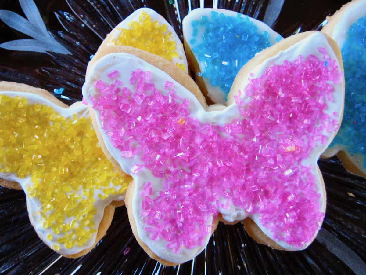Sugar Cookie Cutouts with Royal Icing and pink, yellow and blue decorator sugar shaped like butterflies and served on a crystal plate.