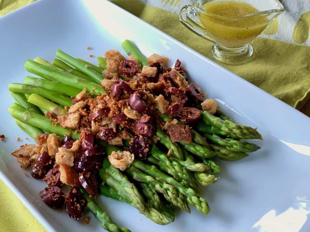 Steamed sparagus topped with toasted crumbs and olives and served on a rectagular platter. A small pitcher of vinaigrette dressing is in the background.