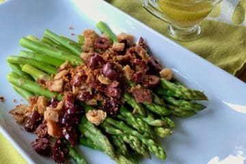 Steamed sparagus topped with toasted crumbs and olives and served on a rectagular platter. A small pitcher of vinaigrette dressing is in the background.