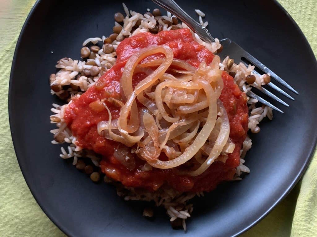 A serving of Kusherie, Egyptian rice and lentils topped with tomato sauce and browned onions, on a black plate with a fork.