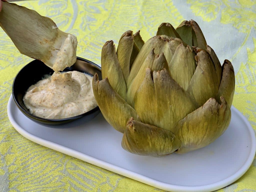 A cooked artichoke on a plate beside one leaf being dipped in a curry dip.