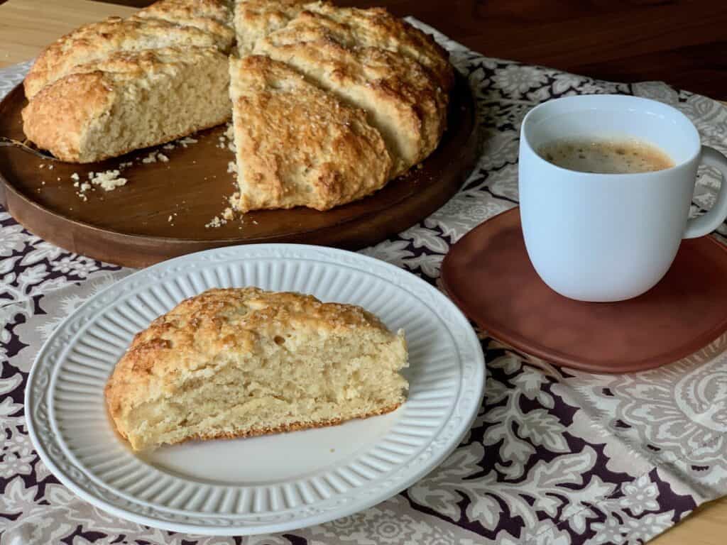 A Buttermilk Scone served on a small plate beside a loaf of scones on a wooden platter and a cup of coffee.