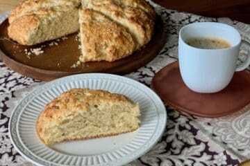 A Buttermilk Scone served on a white plate with more scones in the background and a cup of coffee.