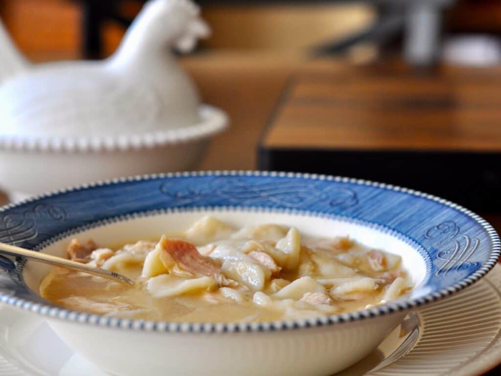 Chicken and Dumplings in a Courier and Ives Ironstone Bowl with a white covered dish shaped like a nesting chicken in the background.