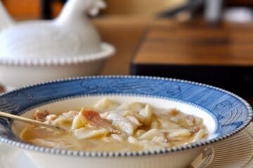 Chicken and Dumplings in a Courier and Ives Ironstone Bowl with a white covered dish shaped like a nesting chicken in the background.