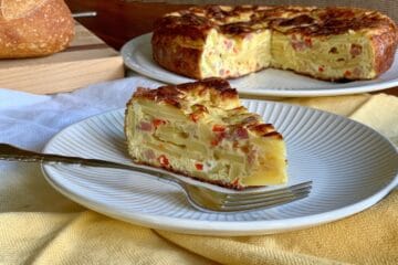 A slice of Spanish Omelet (Tortilla Española) on a white plate with a whole Spanish Omelet and loaf of bread in the background.