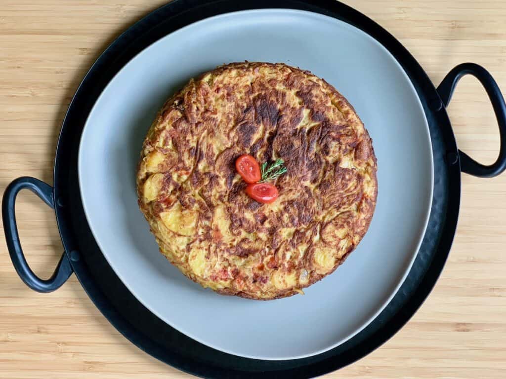 A whole Tortilla Española, or Spanish Omelet, garnished with cherry tomato and rosemary sprig, served on a green platter atop a dark metal tray.