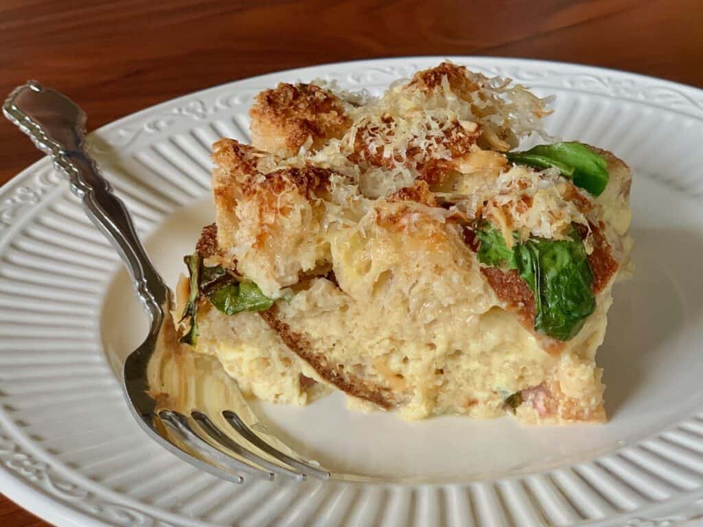 A slice of Arugula and Ham Strata, topped with grated Gruyere, served on a small white plate with a fork.