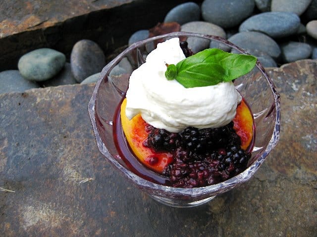 Summer Berry Basil Sauce over a peach half topped with whipped cream and basil leaf in a Glass dish on stone