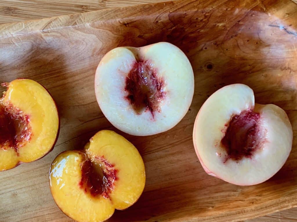 A white and a yellow peach, halved, pitted and displayed on a wood serving dish.