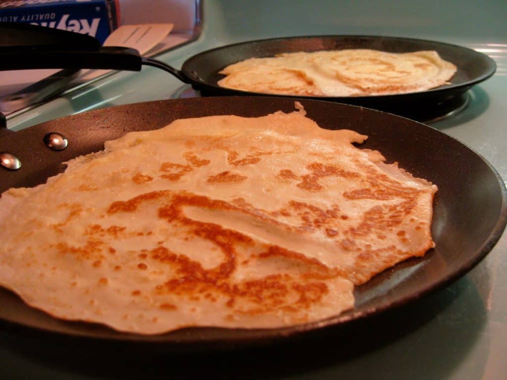 Swedish pancakes being cooked in two flat skillets on a vintage aqua colored cooktop.