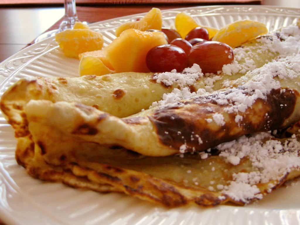 Swedish Pancakes, rolled with Marionberry Jam inside, sprinkled with powdered sugar and served on a plate with a fruit salad.