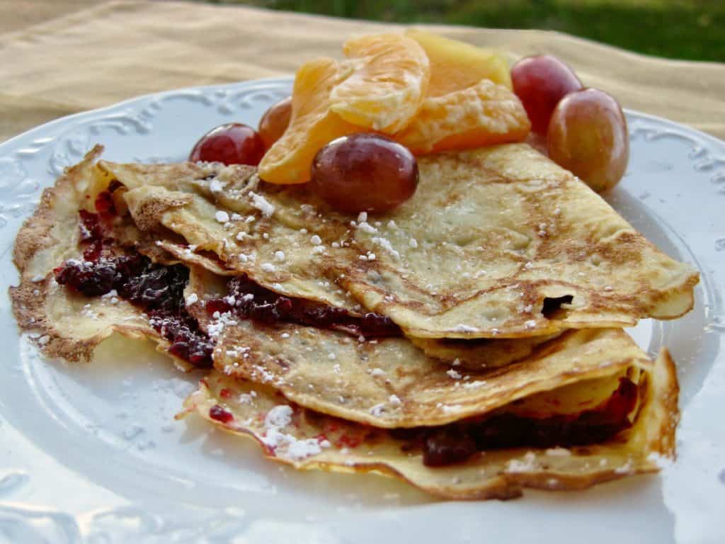 A Swedish Pancake folded with jam inside and sprinkled with powdered sugar. A fruit salad of mandarin sections and grapes is served on the side. 