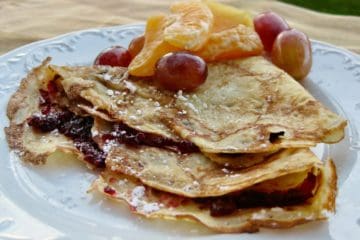 A Swedish Pancake folded with jam inside and sprinkled with powdered sugar. A fruit salad of mandarin sections and grapes is served on the side.