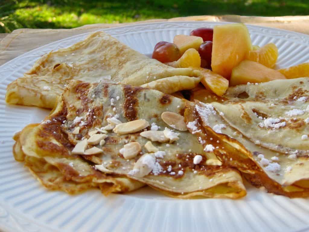 Three Swedish Pancakes folded with jam and sprinkled with toasted almond slices and powdered sugar. A fruit salad of mandarin sections and grapes is served on the side. 
