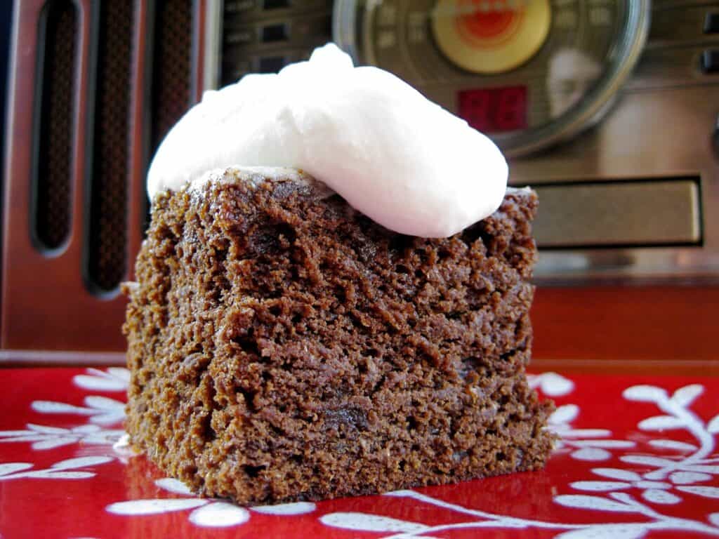 A square of Old-Fashioned Gingerbread topped with Old-Timey Whipped Cream Topping served on a red and white cake.