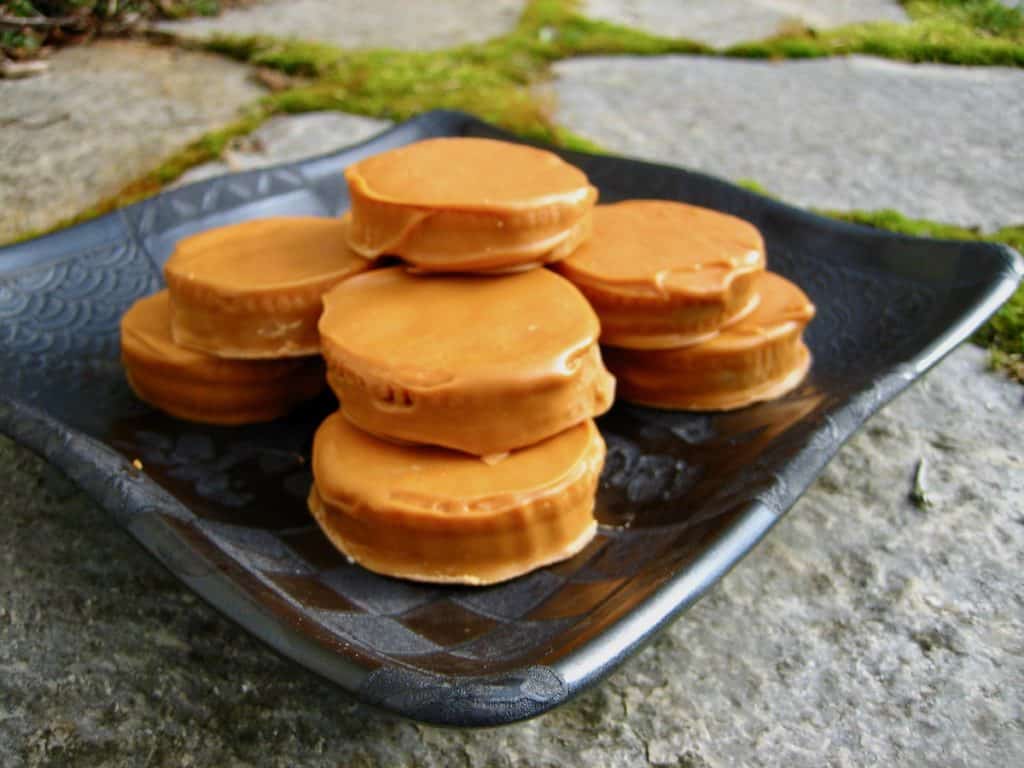 Butterscotch coated Golden Treasure Cookies with peanut butter filling are stacked on a square black plate and displayed on a stone pavement.