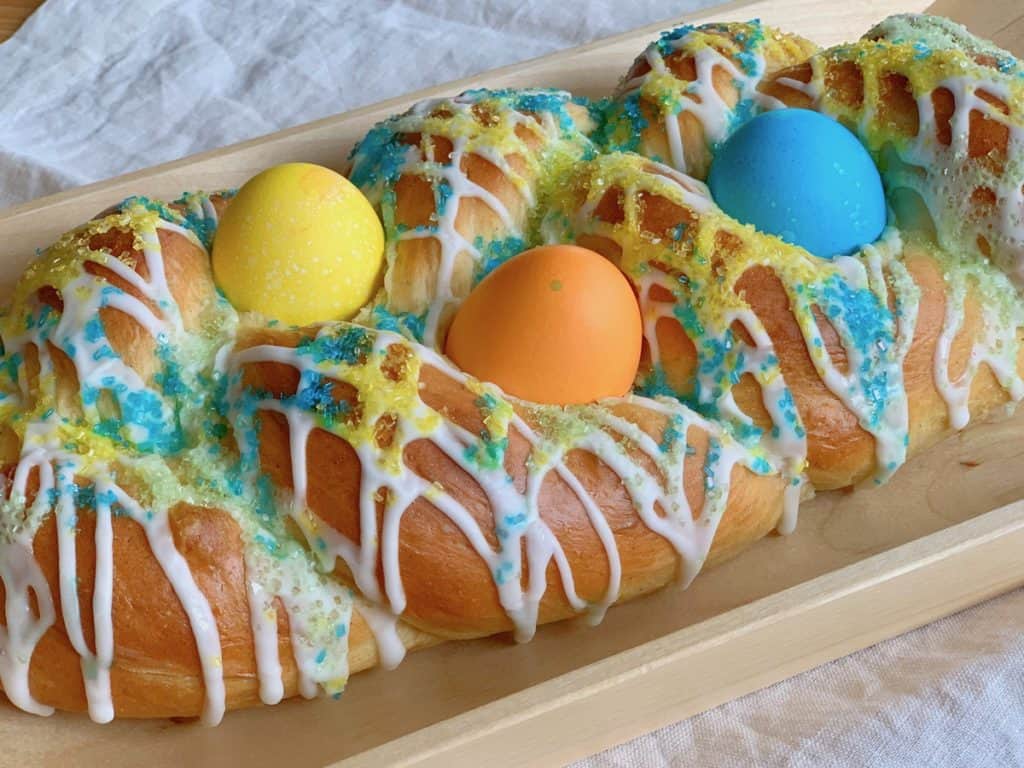 Side view of a Cardamom Citrus Easter Braid drizzled with Lemon Icing, sprinkled with colored sugar and decorated with hard boiled Easter eggs.