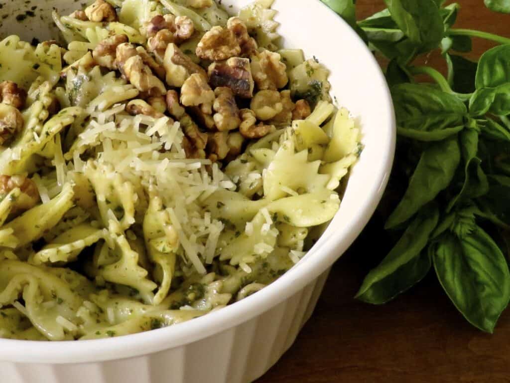 Walnut Pesto on Farfalle, or Bow Tie Pasta, topped with toasted walnuts and grated Parmesan served in a white dish beside a bunch of fresh basil.