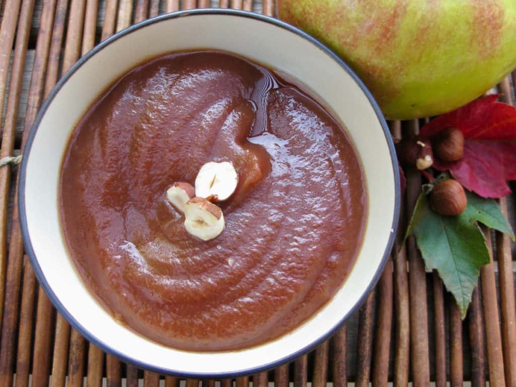 Apple Butter swirled in a bowl topped with a sliced hazelnut set beside autumn apples and leaves.