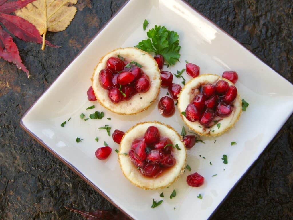 Tiny Goat Cheese Tarts are topped with pomegranate arils in a syrup and arranged on a square plate surrounded by autumn leaves.