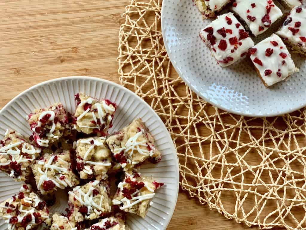 Cranberry Pecan Cookies with White Chocolate Drizzle and Cranberry Bits beside Cranberry Pecan Bars with Orange Cream Cheese Frosting served on white plates and displayed on a woven mat.