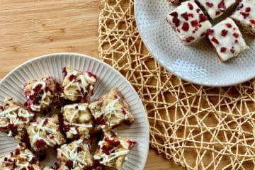 Cranberry Pecan Cookies with White Chocolate Drizzle and Cranberry Bits beside Cranberry Pecan Bars with Orange Cream Cheese Frosting served on white plates and displayed on a woven mat.