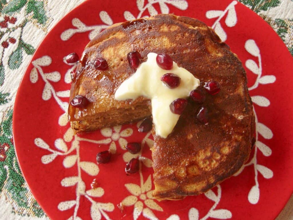 A cut stack of Gingerbread Pancakes topped with butter, syrup and pomegranate arils on a festive red and white plate.