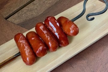 Cocktail Wieners in Bourbon Sauce Skewered on an iron serving pic and resting on a wooden board.