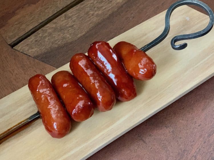 Cocktail Wieners in Bourbon Sauce Skewered on an iron serving pic and resting on a wooden board.