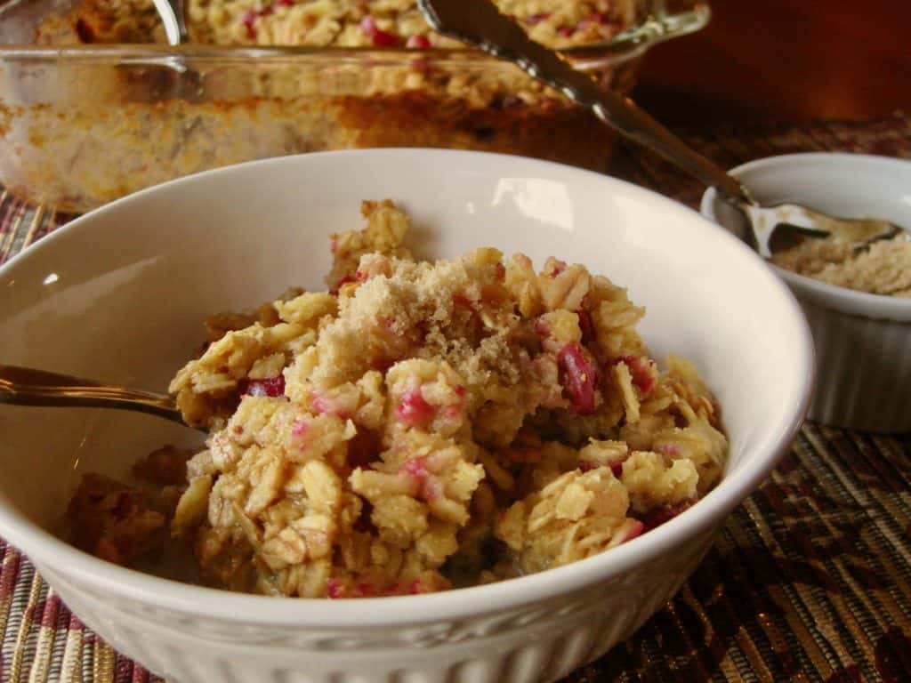 A bowl filled with Cranberry Orange Baked Oatmeal with the baked oatmeal casserole and a dish of brown sugar in the background.