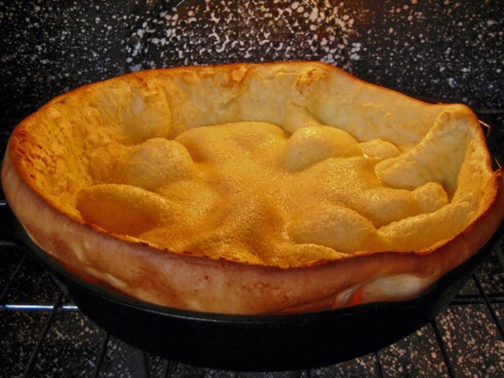 A Dutch Baby Pancake in the oven is puffed and arching over the sides of an iron skillet.