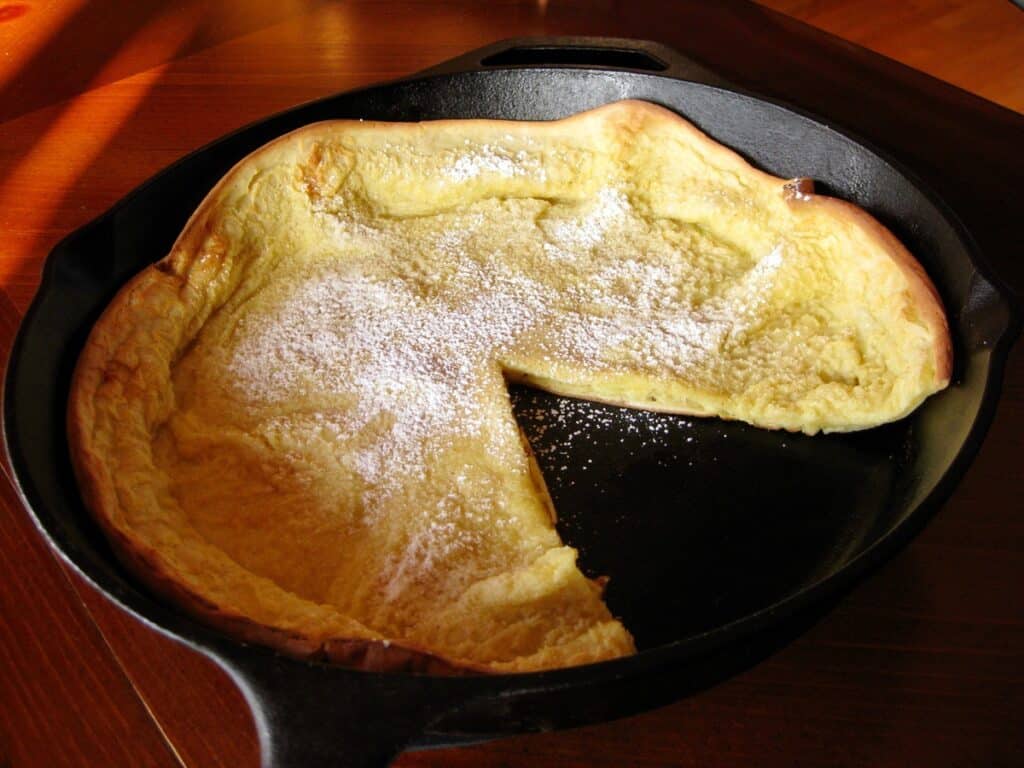 Dutch Baby Pancake in Iron Skillet, dusted with powder sugar, has one slice removed.