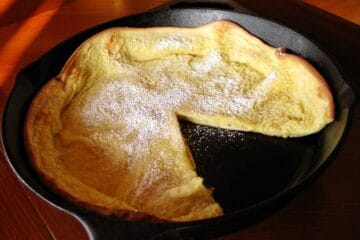 Dutch Baby Pancake in Iron Skillet, dusted with powder sugar, has one slice removed.