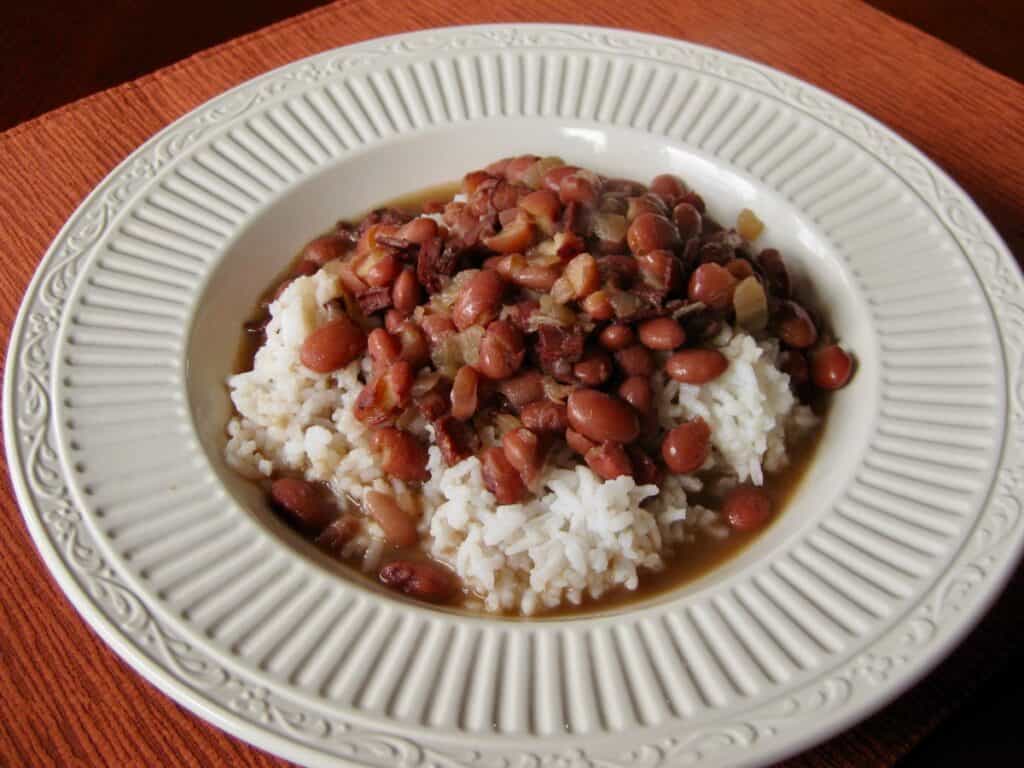 Red Beans, seasoned with cinnamon, bay leaves and ham, is served over rice in a shallow white bowl.
