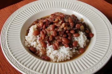Red Beans, seasoned with cinnamon, bay leaves and ham, is served over rice in a shallow white bowl.