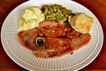 A Slice of Kentucky Country Ham covered with Red Eye Gravy served on a dinner plate beside Southern Green Beans, Mashed Potatoes and a sliced Biscuit.
