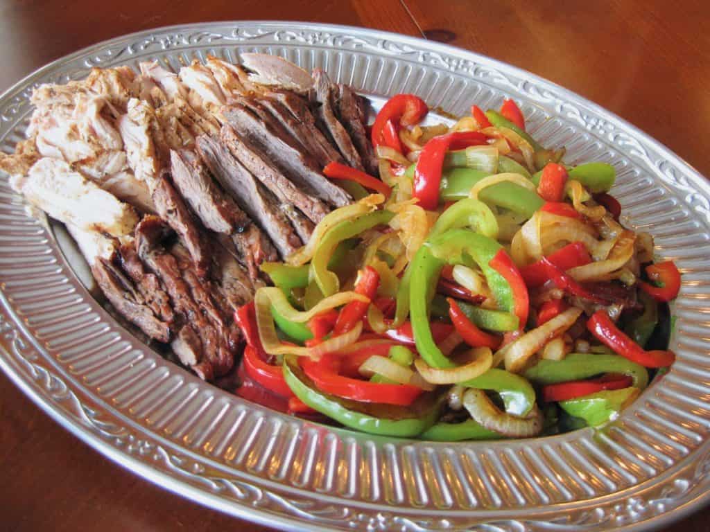 Sliced red and green bell peppers sautéed with onions are arranged on a platter with beef and chicken to make fajitas.