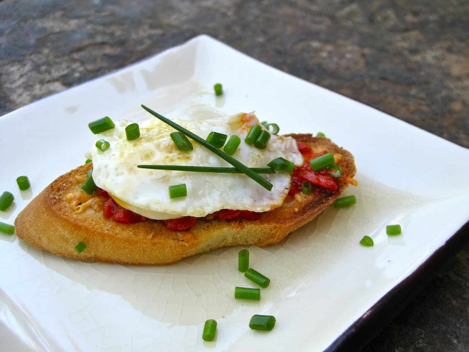 A pretty Bruschetta made from Crostini topped with red pepper sauce and a fried quail egg sprinkled with fresh herbs.